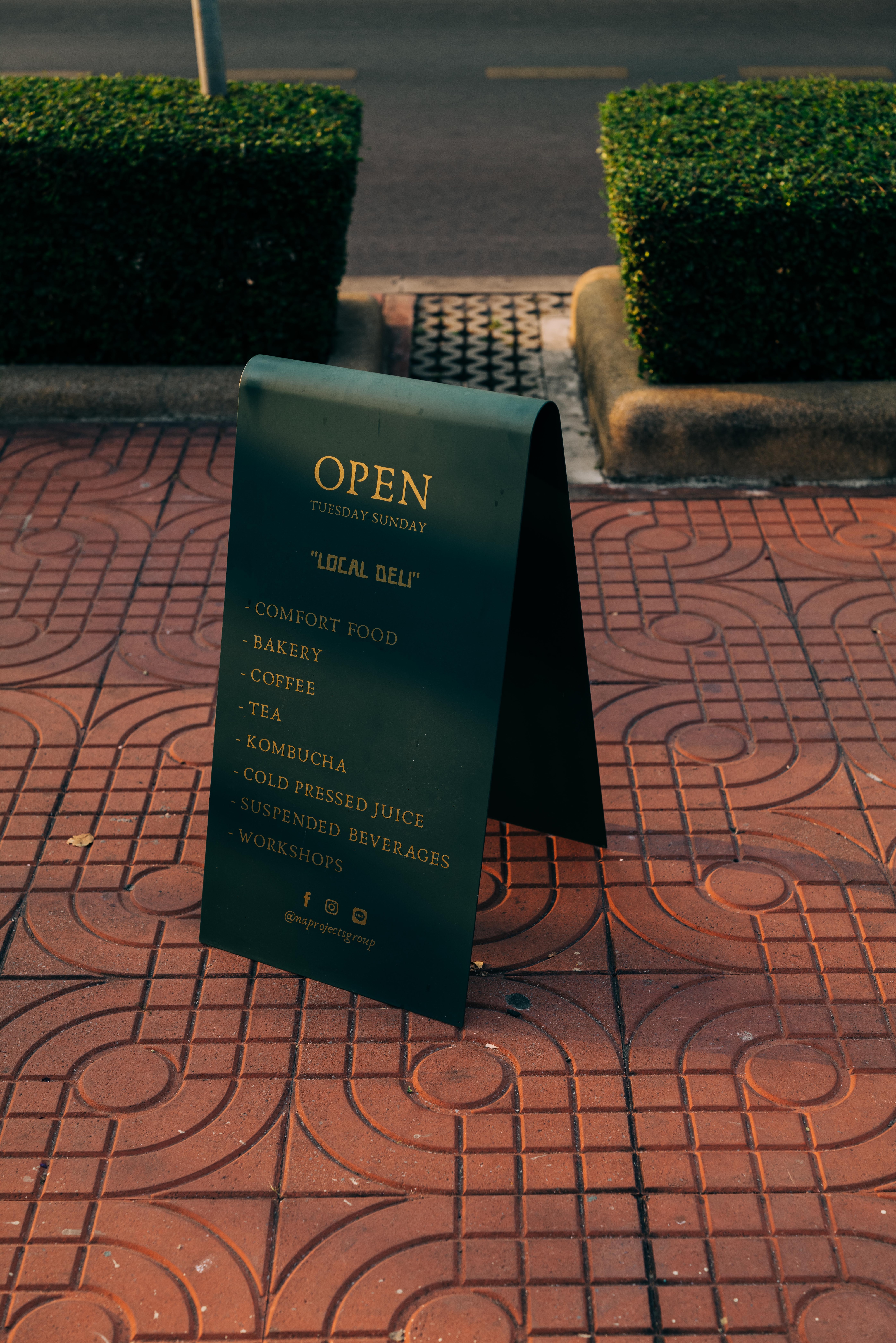 A dark green sign on orange-red colored stones reads "Open - Local Deli" with a list of food and beverage items.